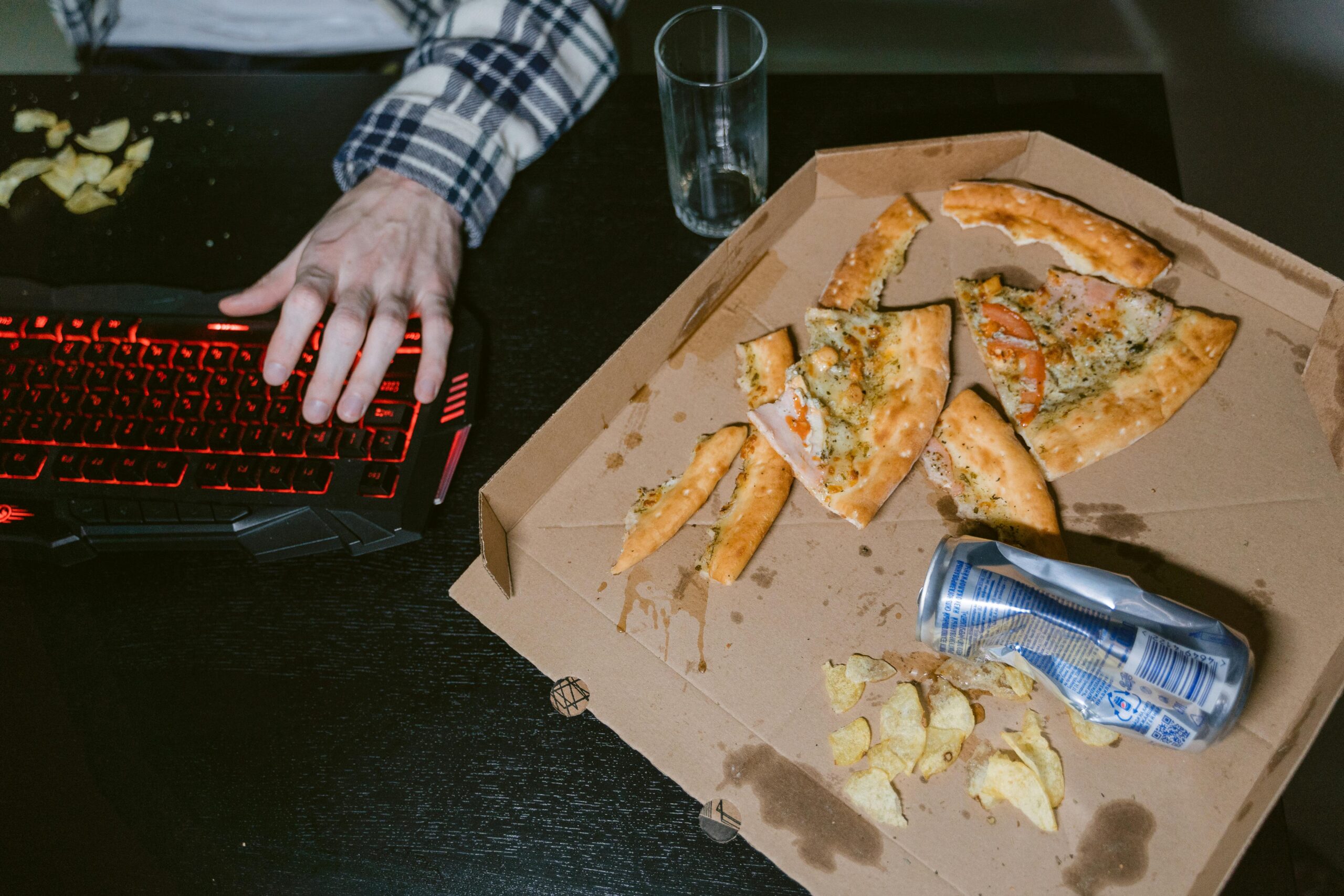A gamer enjoys pizza and soda in a dimly lit room with a red-lit keyboard.